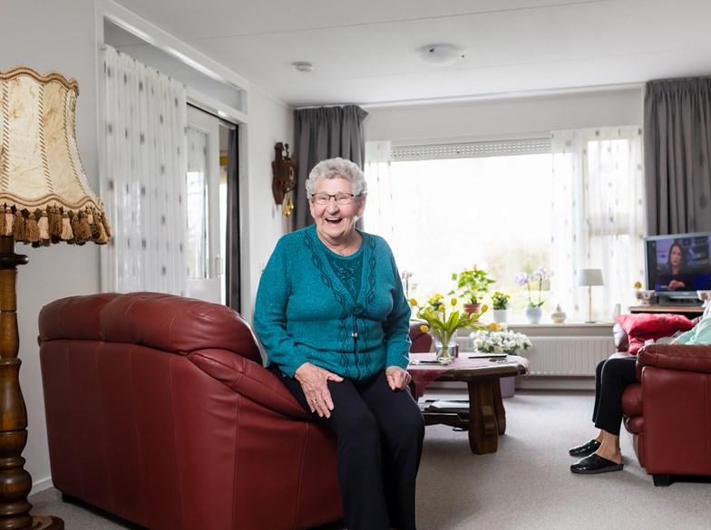 Oudere vrouw kijkt vanuit haar huiskamer lachend op een leren stoel in camera